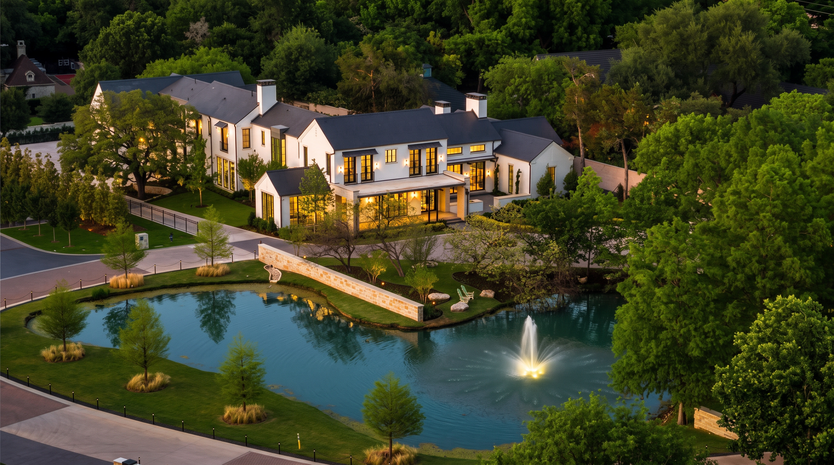 This high-angle aerial view captures a sprawling, modern estate nestled within a lush, tree-filled landscape. The property features a white-painted home with a dark roof, a private pond with a central fountain, and manicured green lawns. The perspective emphasizes the expansive grounds and the serene, park-like setting of the residence.