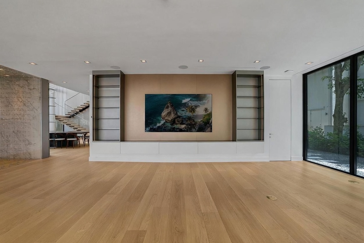 This spacious, minimalist living room features light wood flooring and a central media wall with a large television mounted against a metallic-toned panel. Flanking the television are built-in shelving units with integrated lighting, set above a long, low-profile white storage console. To the right, large glass panels provide a view of an outdoor garden, while the left side opens into a dining area with a staircase visible in the background.