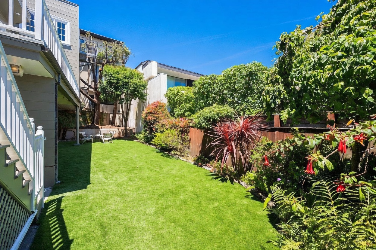 This backyard features a well-maintained artificial turf lawn bordered by a wooden fence and lush, mature landscaping. A white staircase descends from the side of the house, leading to a small seating area tucked near the back of the property. The scene is captured from a ground-level perspective, highlighting the private, sun-drenched outdoor space.