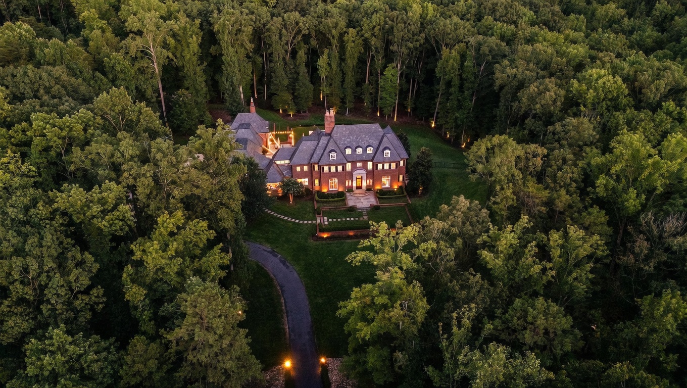 This high-angle aerial shot captures a grand, multi-story brick estate nestled within a dense, lush forest. The home features a complex roofline with multiple gables, a paved driveway winding through the trees, and a manicured lawn with structured landscaping near the entrance. The warm glow from the interior lights and exterior accent lighting creates an inviting and prestigious atmosphere at dusk.