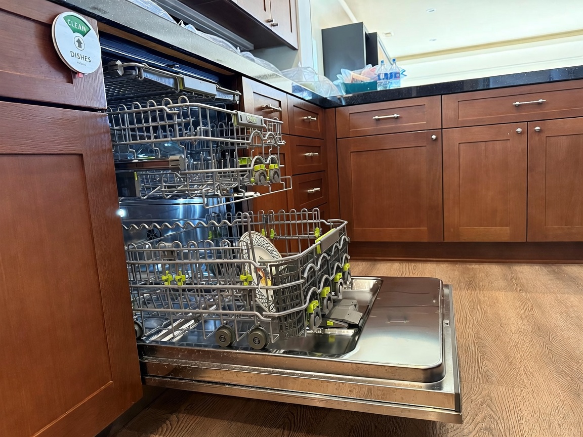 This image provides a close-up, eye-level perspective of an open stainless steel dishwasher integrated into dark wood cabinetry. The dishwasher racks are extended, revealing a clean interior, while a 'Clean Dishes' magnet is attached to the adjacent cabinet door. The scene highlights the kitchen's functional layout, featuring dark wood-toned cabinets and a dark countertop surface.