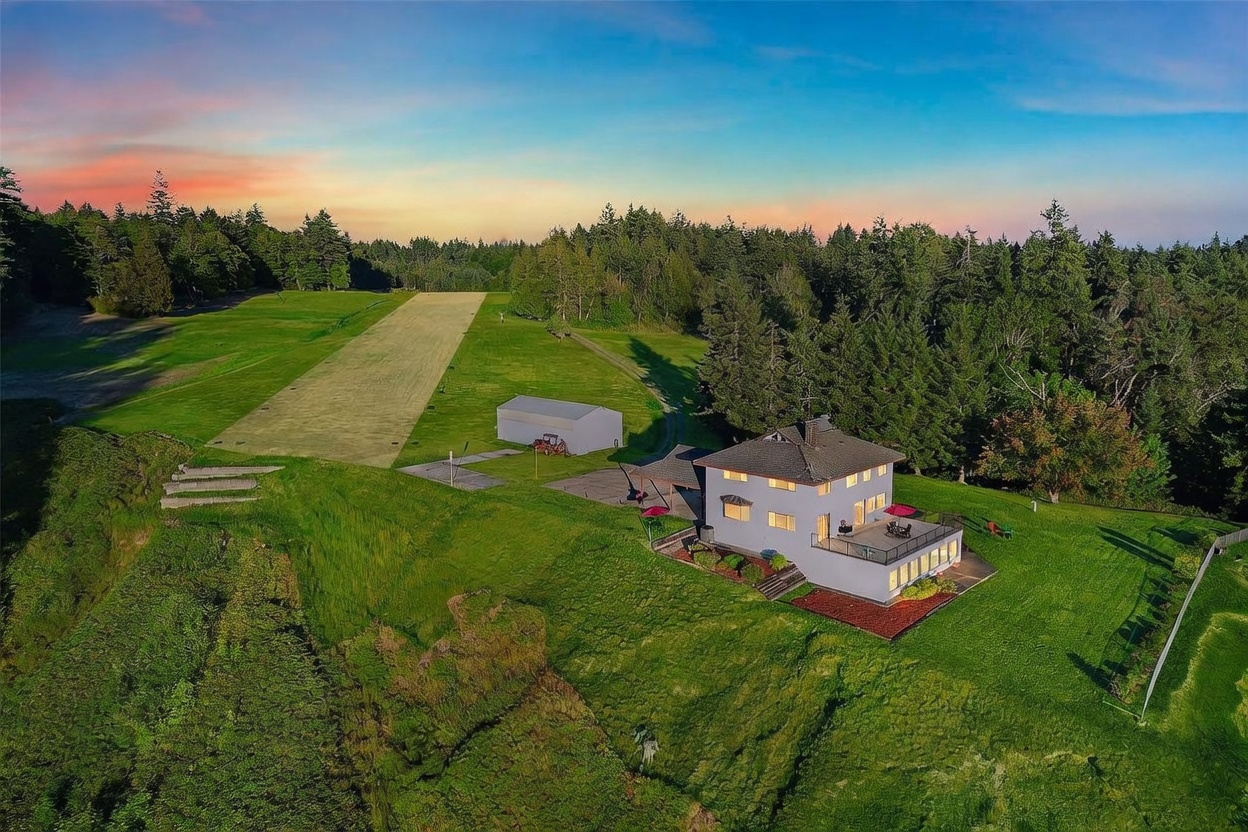 This high-angle aerial view captures a multi-story home situated on a sprawling, grassy property that includes a private landing strip. The house features a large, elevated deck overlooking the expansive lawn and surrounding dense forest, with a detached metal outbuilding nearby. The scene is bathed in the warm, golden light of a sunset, emphasizing the property's secluded and unique rural character.