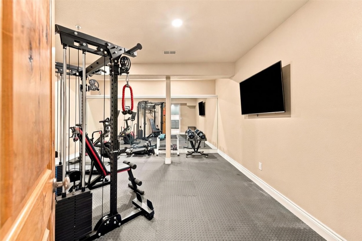 This dedicated home gym features a functional cable machine, a weight bench, and a rack of dumbbells, all set upon durable, textured rubber flooring. A large wall-mounted television provides entertainment, while a full-width mirror reflects the equipment, creating an expansive feel in the space. The room is finished with neutral-toned walls and recessed lighting, offering a clean and practical environment for fitness.