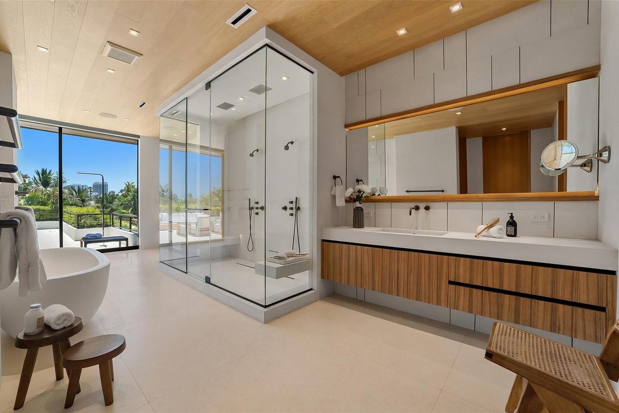 This luxurious primary bathroom features a minimalist aesthetic with a large, glass-enclosed walk-in shower and a freestanding soaking tub positioned near floor-to-ceiling windows that offer a view of the outdoors. A long, wall-mounted vanity with light wood cabinetry and a white countertop provides ample space, complemented by a wide mirror and modern fixtures. The room is bathed in natural light, highlighting the clean lines and the warm, light-toned wood ceiling.