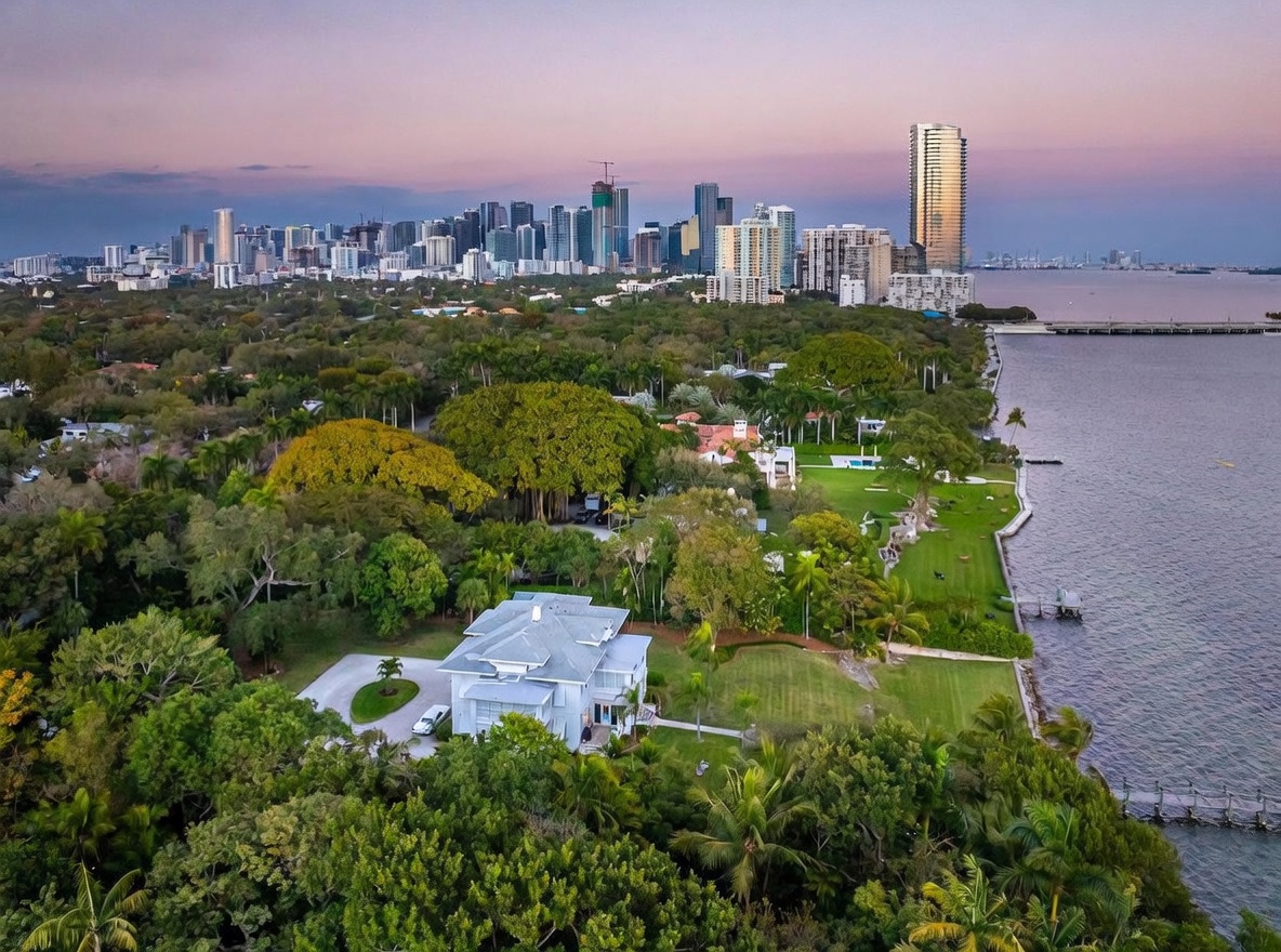 This high-angle aerial shot captures a sprawling waterfront estate nestled among lush, mature trees along a bay. The property features a large, light-colored residence with a multi-pitched roof, expansive green lawns, and a private dock extending into the water. In the background, a vibrant city skyline glows under a soft sunset sky, providing a striking contrast between the serene residential landscape and the urban horizon.