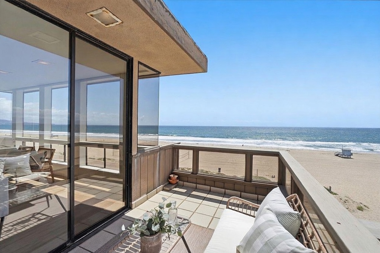 This elevated balcony offers a direct, panoramic view of the ocean and sandy beach, featuring a glass-railed deck furnished with comfortable outdoor seating. Large sliding glass doors provide seamless indoor-outdoor flow, reflecting the bright, sunny coastal environment. The space is designed for relaxation, capturing the essence of beachfront living with its unobstructed horizon and tranquil atmosphere.