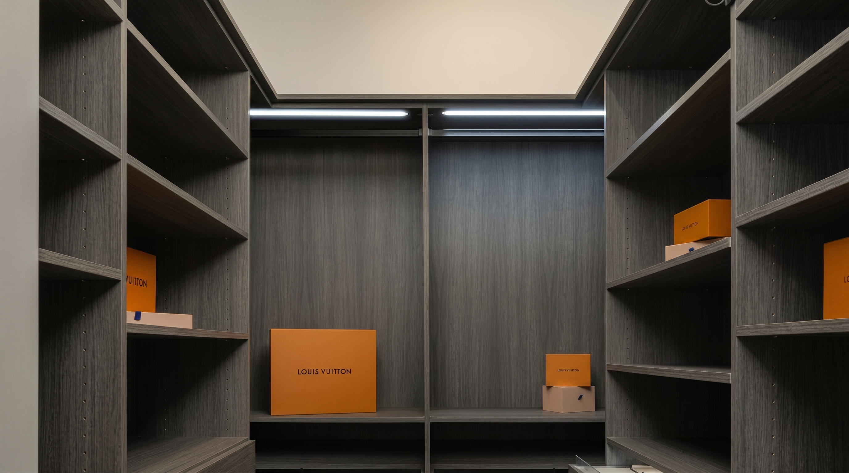 This walk-in closet is characterized by custom, dark wood cabinetry with integrated shelf lighting and a U-shaped layout that maximizes storage space. The design is sleek and contemporary, providing an organized display area, currently accented with luxury Louis Vuitton packaging. The clean lines and minimalist aesthetic create an upscale feel suited for a high-end property.