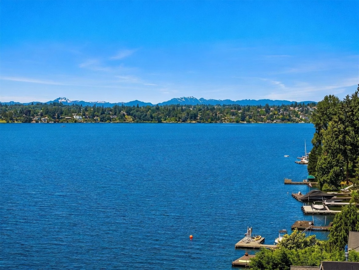 This scenic exterior view captures a expansive, deep blue lake framed by lush green trees and distant, snow-capped mountains under a clear sky. In the foreground, several private wooden docks extend into the water, with a small boat moored at one, emphasizing the waterfront lifestyle. The perspective is elevated, likely from a balcony or deck, providing a panoramic and tranquil outlook over the property's natural surroundings.