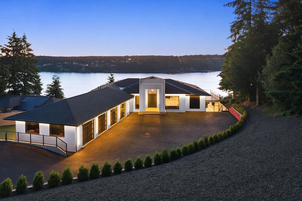 This elevated perspective captures a modern, single-story residence at dusk, set against a serene waterfront backdrop. The home features a prominent, central entryway with a tall, rectangular architectural frame, flanked by a multi-car garage on the left and expansive living areas on the right. A paved driveway leads to the entrance, bordered by a neat row of small evergreen shrubs and a gravel path, creating a clean and sophisticated curb appeal.
