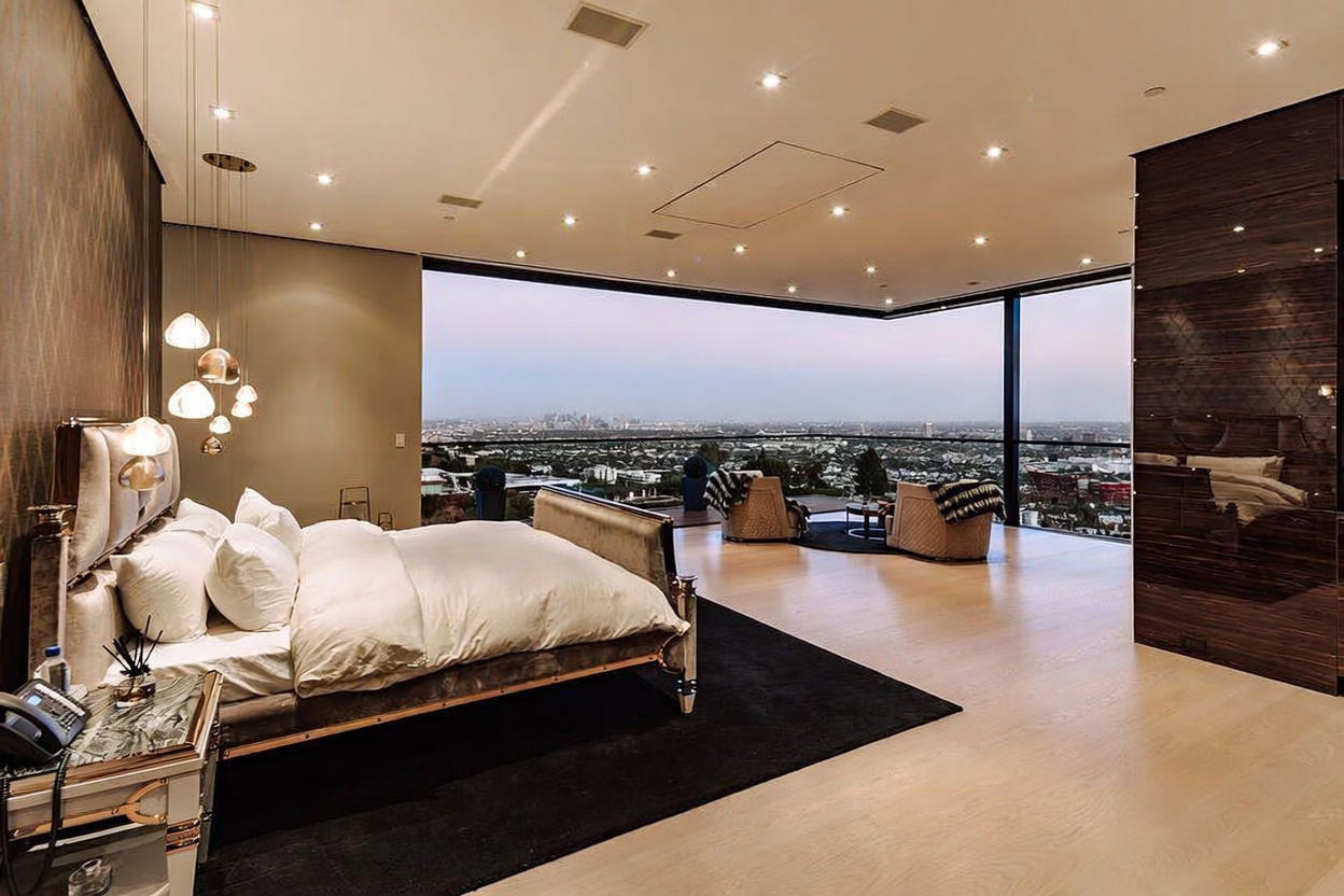 This luxurious primary bedroom features a contemporary design with a plush bed, elegant pendant lighting, and a dark accent wall. The room is defined by its expansive floor-to-ceiling glass walls that offer a breathtaking, panoramic view of the city skyline. The open-concept layout includes a cozy seating area, creating a sophisticated and serene atmosphere.