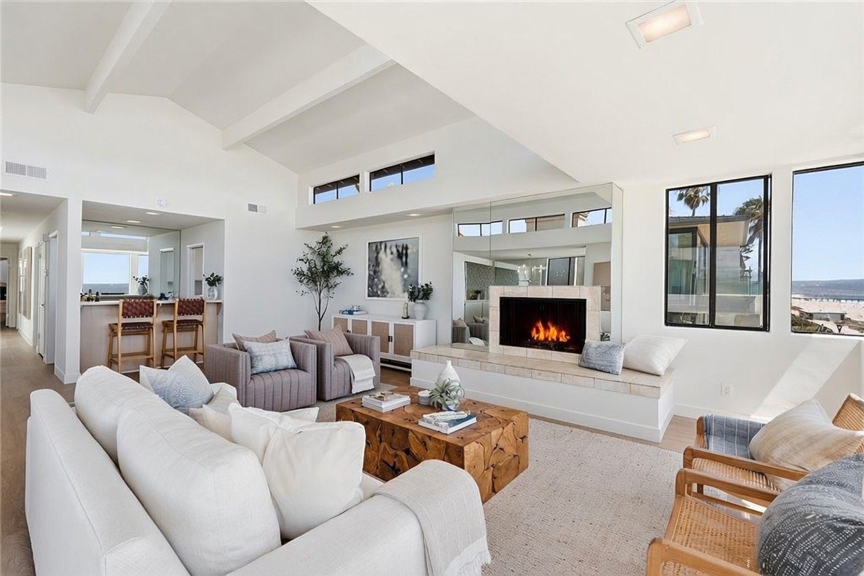 This bright and airy living room features a vaulted ceiling with exposed beams and a neutral color palette that emphasizes a relaxed, coastal aesthetic. A central fireplace with a tiled hearth serves as a focal point, complemented by a large mirror above it and comfortable seating arranged around a rustic wooden coffee table. Large windows provide ample natural light and offer a scenic view of the beach and ocean in the distance.