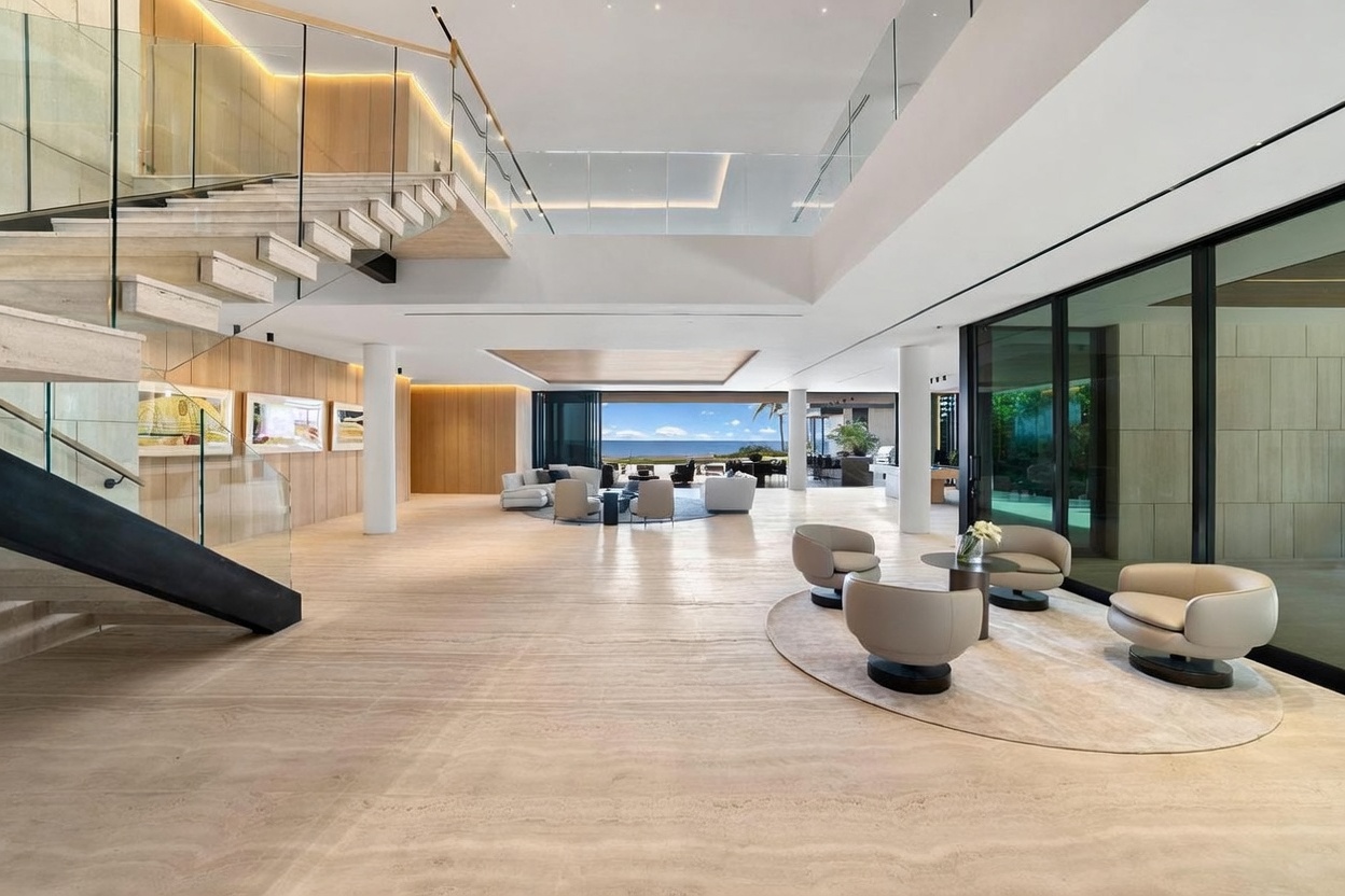 This expansive, open-concept living area features a grand floating staircase with glass railings and light-toned stone flooring throughout. The space is anchored by two distinct seating arrangements, including a circular rug with modern armchairs in the foreground and a larger lounge area further back that opens directly to an ocean view through expansive glass walls. The minimalist design emphasizes volume and light, creating a sophisticated and airy atmosphere suitable for luxury coastal living.