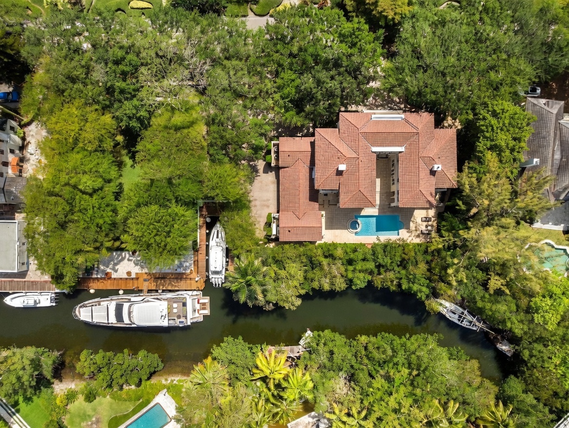 This high-angle aerial view captures a sprawling waterfront estate nestled among lush, mature trees. The property features a large residence with a terracotta-tiled roof, a private swimming pool, and a dedicated boat slip housing a yacht along a narrow canal. The surrounding landscape is densely wooded, providing a secluded and tranquil atmosphere for this luxury waterfront home.