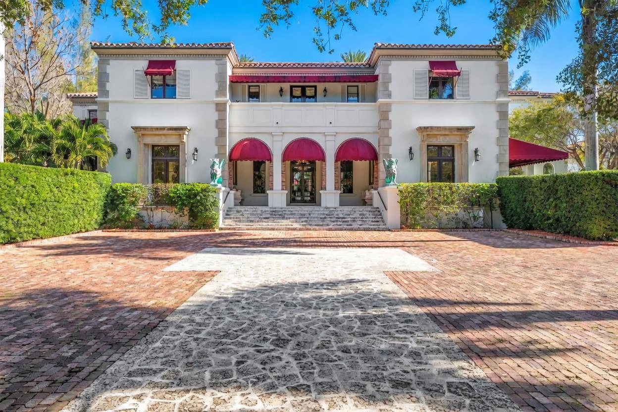 This grand Mediterranean-style estate features a symmetrical white stucco facade with prominent red fabric awnings over the arched entryway and upper-level windows. A wide, stone-paved driveway leads to a set of central stairs, flanked by lush green hedges and decorative statues. The perspective is a straight-on, eye-level shot that emphasizes the property's imposing scale and formal architectural design.