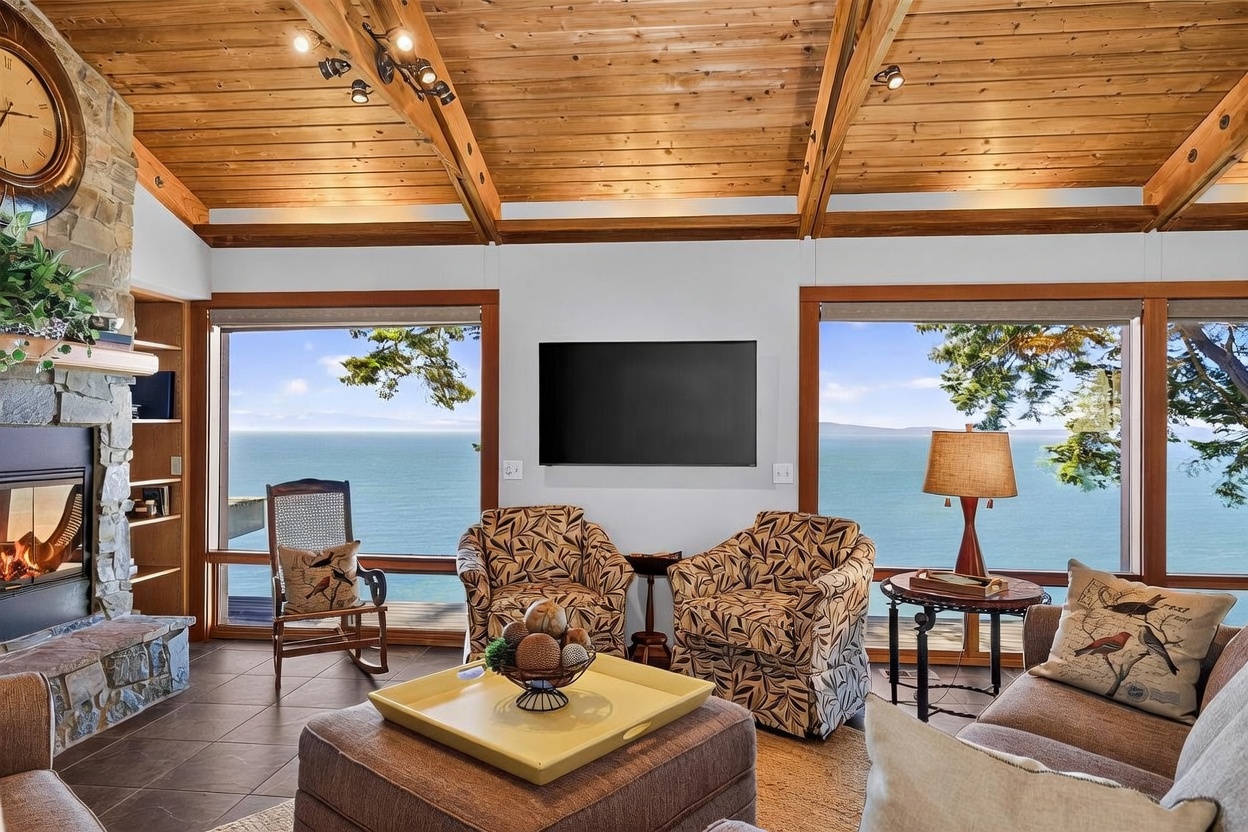 This inviting living room features a vaulted wood-paneled ceiling with exposed beams and large windows that frame a panoramic ocean view. A stone-clad chimney breast anchors the space, complemented by a cozy seating area with patterned armchairs, a rocking chair, and a central ottoman. The room exudes a warm, rustic atmosphere, perfectly blending comfortable indoor living with the natural beauty of the coastal landscape.