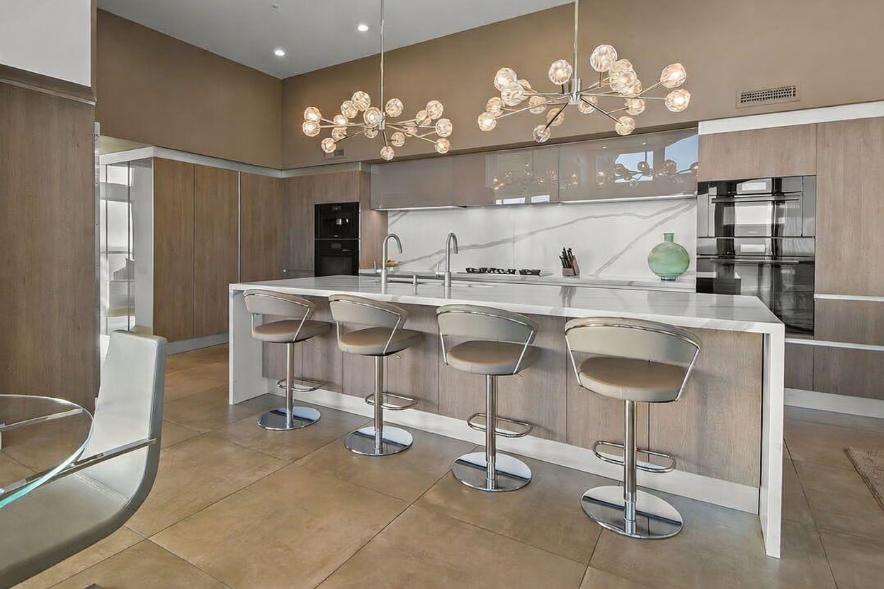 This modern kitchen features a large central island with a stone countertop and four bar stools, serving as the focal point of the room. The space is accented by two intricate, multi-bulb pendant light fixtures and sleek, light-toned wood cabinetry paired with glossy upper cabinets. A clean, minimalist aesthetic is maintained through the integrated appliances and a light-colored stone backsplash, creating a sophisticated and airy atmosphere.