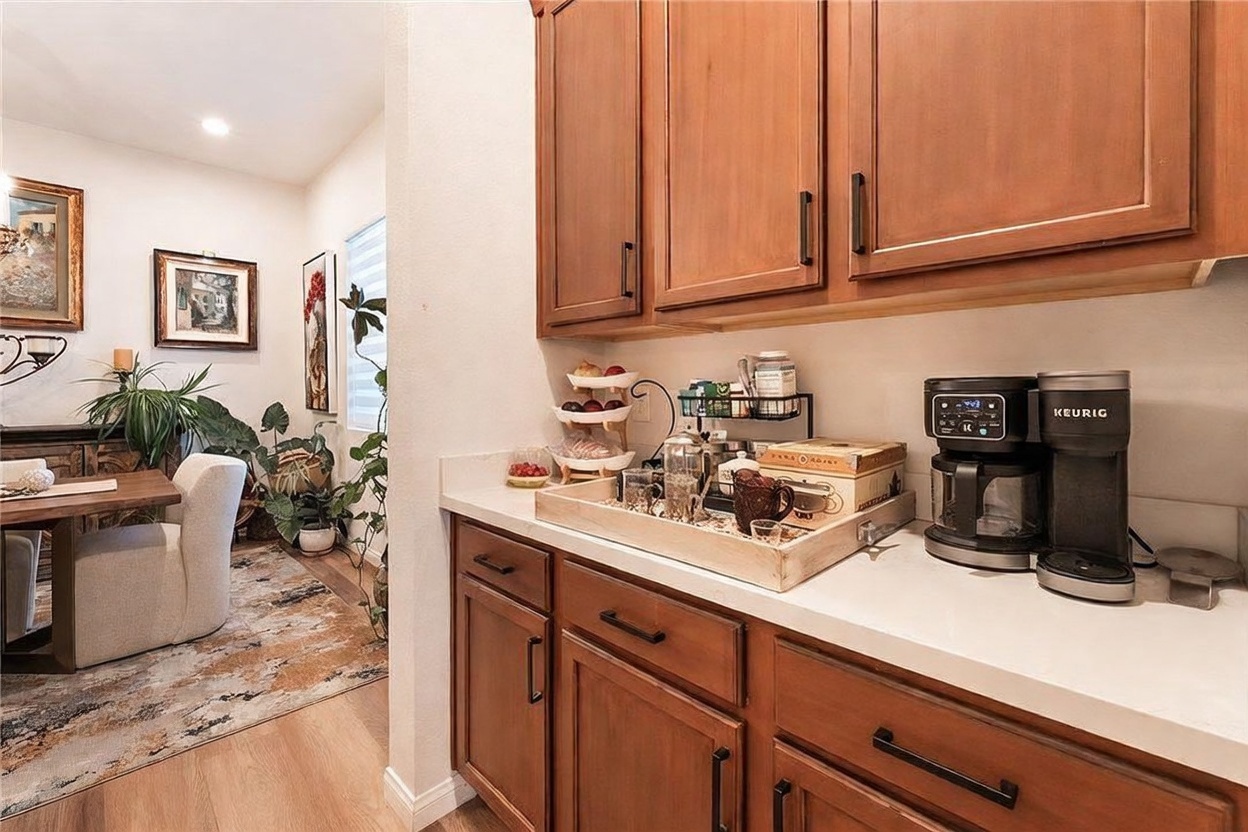 This image captures a functional coffee station area within a kitchen, featuring warm-toned wood cabinetry and white countertops. A wooden tray organizes various coffee supplies and a Keurig machine, while an adjacent dining area is visible through the open transition, showcasing a dining table, chairs, and framed wall art. The perspective is a medium shot from the side, highlighting the seamless flow between the kitchen workspace and the dining space.