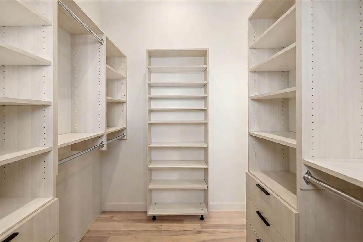 This walk-in closet features light wood-toned cabinetry with extensive shelving, hanging rods, and built-in drawers. A central, mobile shelving unit provides additional storage, while the light-colored walls and wood flooring create a bright, organized atmosphere. The symmetrical layout and clean lines offer a modern and functional storage solution.