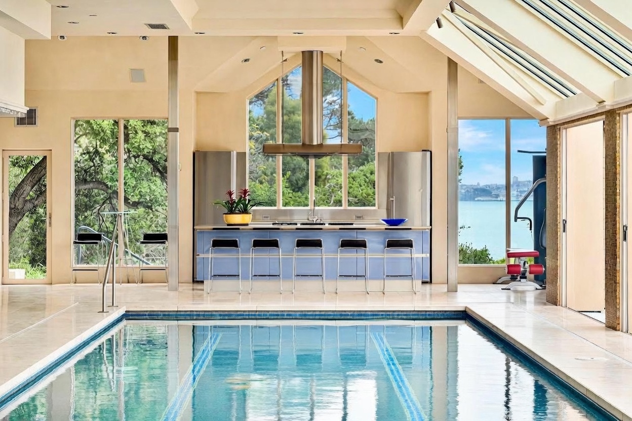 This expansive indoor pool area features a lap pool as the central focus, complemented by a built-in kitchen bar with five stools. The room is bathed in natural light from large windows and a sloped glass ceiling, offering scenic views of the water and distant cityscape. The space combines leisure and entertainment, with a home gym area visible to the right and a sleek, modern aesthetic throughout.