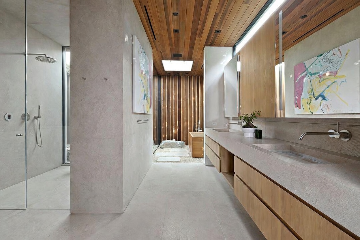 This luxurious, spa-inspired primary bathroom features a minimalist design with a warm wood-paneled ceiling and a long, floating double vanity with integrated stone sinks. A glass-enclosed walk-in shower sits to the left, while the back of the room opens into a serene, zen-like area with a wooden soaking tub and decorative stone accents. The space is bright and airy, utilizing a neutral color palette and modern art to create a sophisticated, high-end atmosphere.