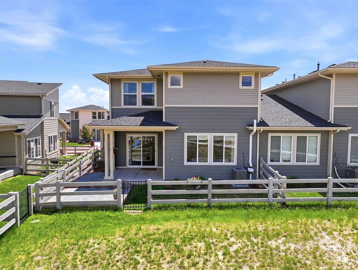 This rear view of a two-story residential property showcases a gray-sided exterior with a paved patio area enclosed by a white picket fence. The home features a mix of horizontal and shingle-style siding, large windows, and a sliding glass door leading to the outdoor living space. The perspective is captured from the backyard, highlighting the private, fenced-in lawn and the clean, modern architectural lines of the building.