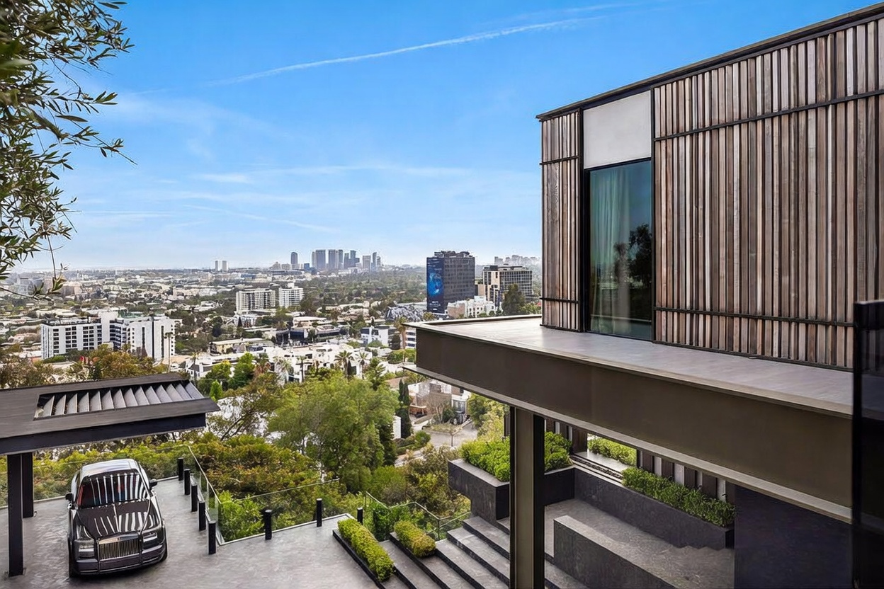 This striking modern architectural home features a sleek, multi-level design with vertical wood slat siding and expansive glass windows that frame a panoramic view of the city skyline. A luxury SUV is parked in the covered driveway area, which transitions into tiered stone steps and landscaped planters leading down the hillside. The perspective captures the seamless integration of high-end contemporary design with the dramatic, elevated landscape.