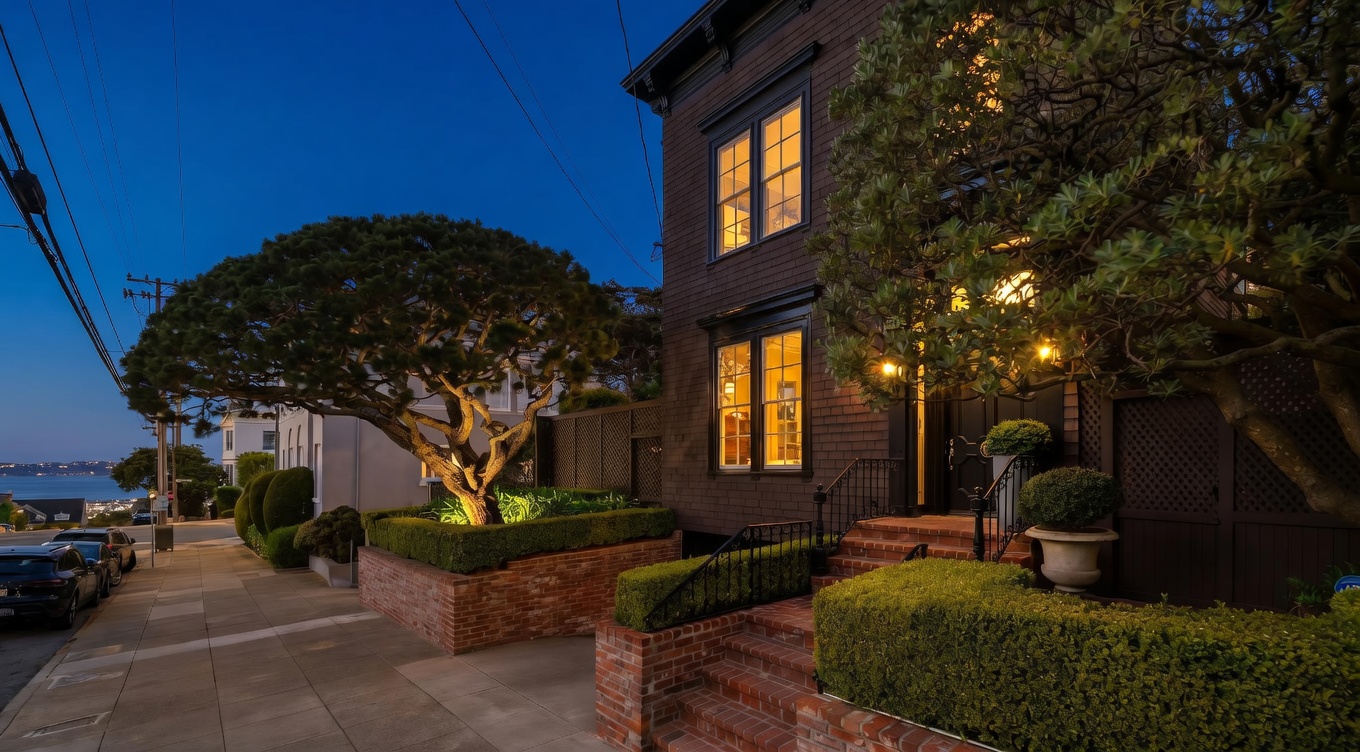 This charming, dark-toned residence features a classic architectural style with a prominent brick-walled garden and manicured hedges lining the front walkway. The perspective captures the home from the sidewalk, showcasing its elevated entryway, tall windows with dark frames, and a mature tree that frames the facade. The setting includes a view of the bay in the distance, contributing to a sophisticated and well-maintained curb appeal.