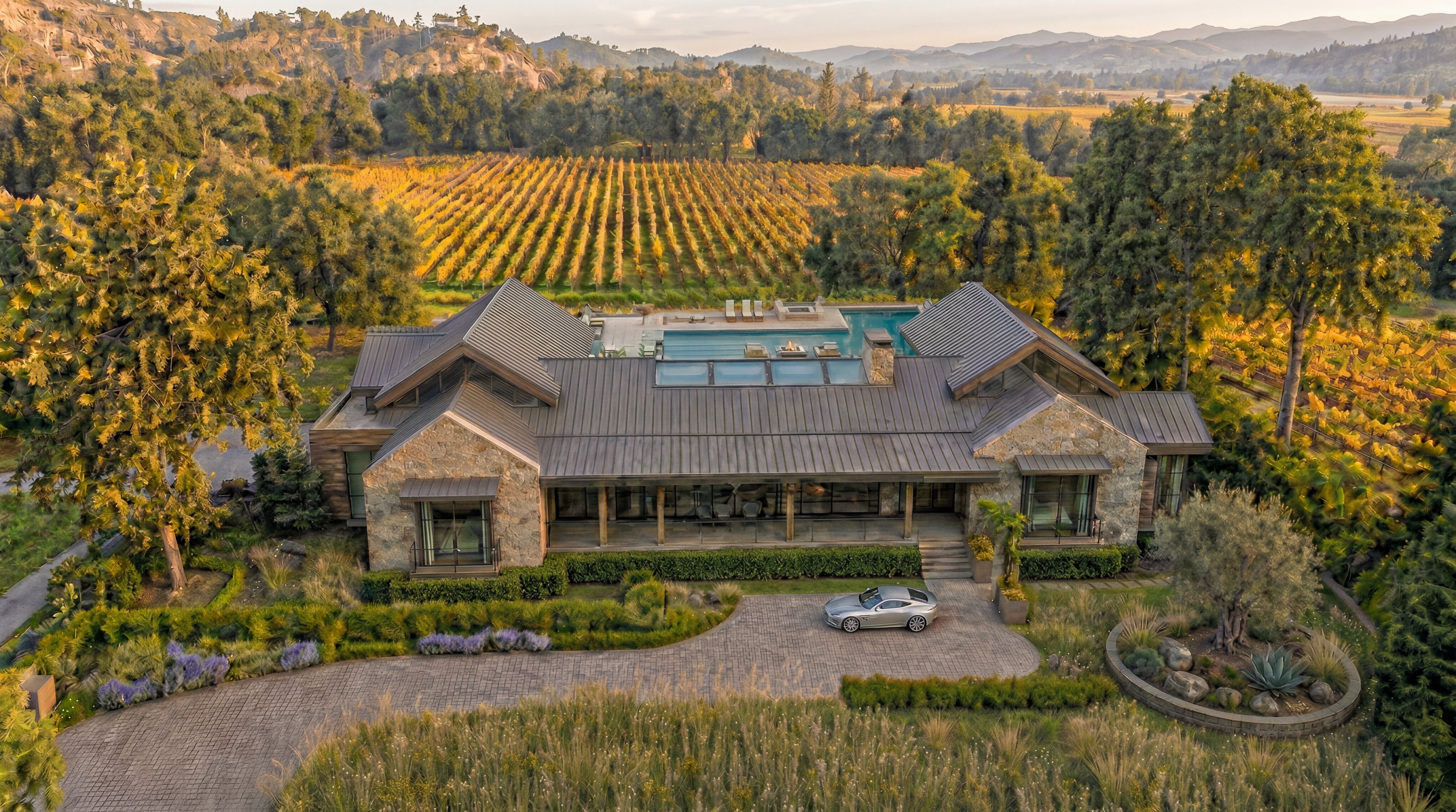 This stunning rural estate features a sophisticated contemporary house built with rustic stone and a metal roof, overlooking a beautifully manicured vineyard. The property is highlighted by a luxury infinity pool on the rear terrace and a paved driveway leading to the front entry, where a luxury car is parked. The overall design blends modern architectural clean lines with the natural beauty and textures of the surrounding countryside.