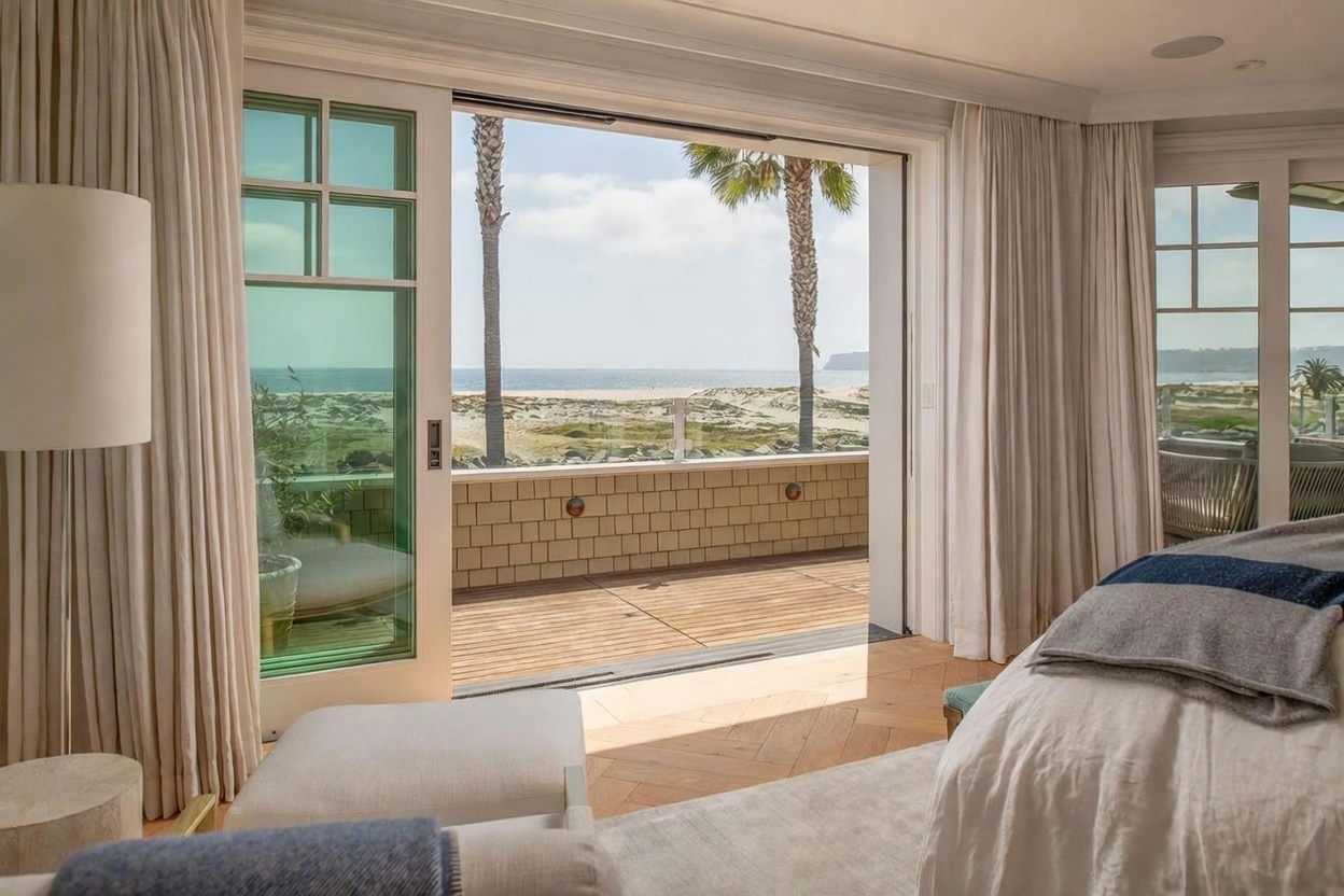This primary bedroom features a seamless transition to an outdoor deck through large sliding glass doors that frame a panoramic ocean view. The interior is finished with light wood flooring in a herringbone pattern and neutral-toned furnishings, creating a serene and airy atmosphere. The perspective captures the inviting indoor-outdoor connection, highlighting the proximity to the beach and the natural light flooding the space.
