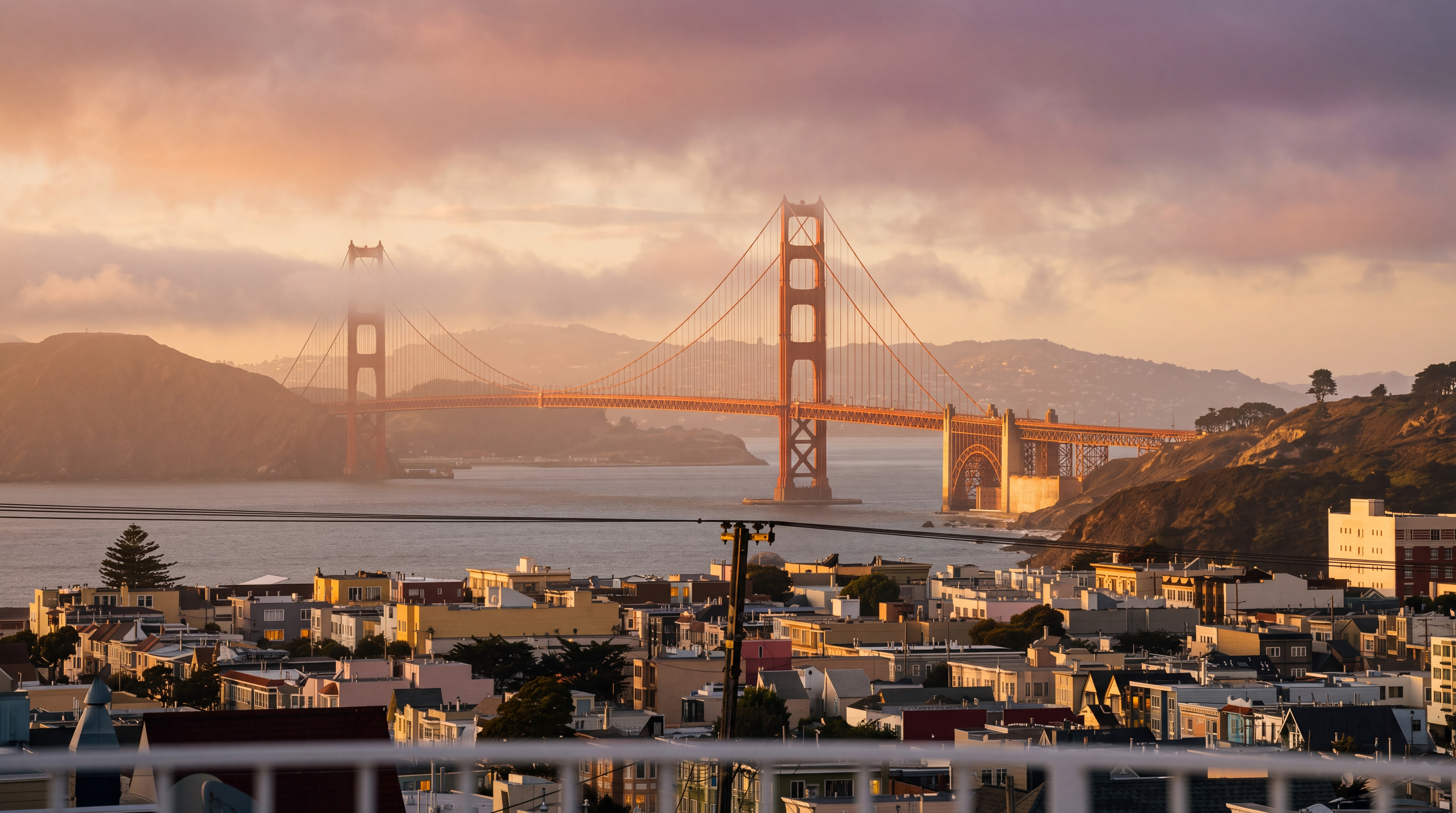 This image is captured from an elevated balcony, offering a breathtaking, panoramic view of the iconic Golden Gate Bridge set against a vibrant sunset sky. In the foreground, a white railing frames the scene, looking out over a dense urban neighborhood of San Francisco homes nestled on the hillsides. The perspective provides a cinematic sense of scale, emphasizing the property's prime location and spectacular vistas.