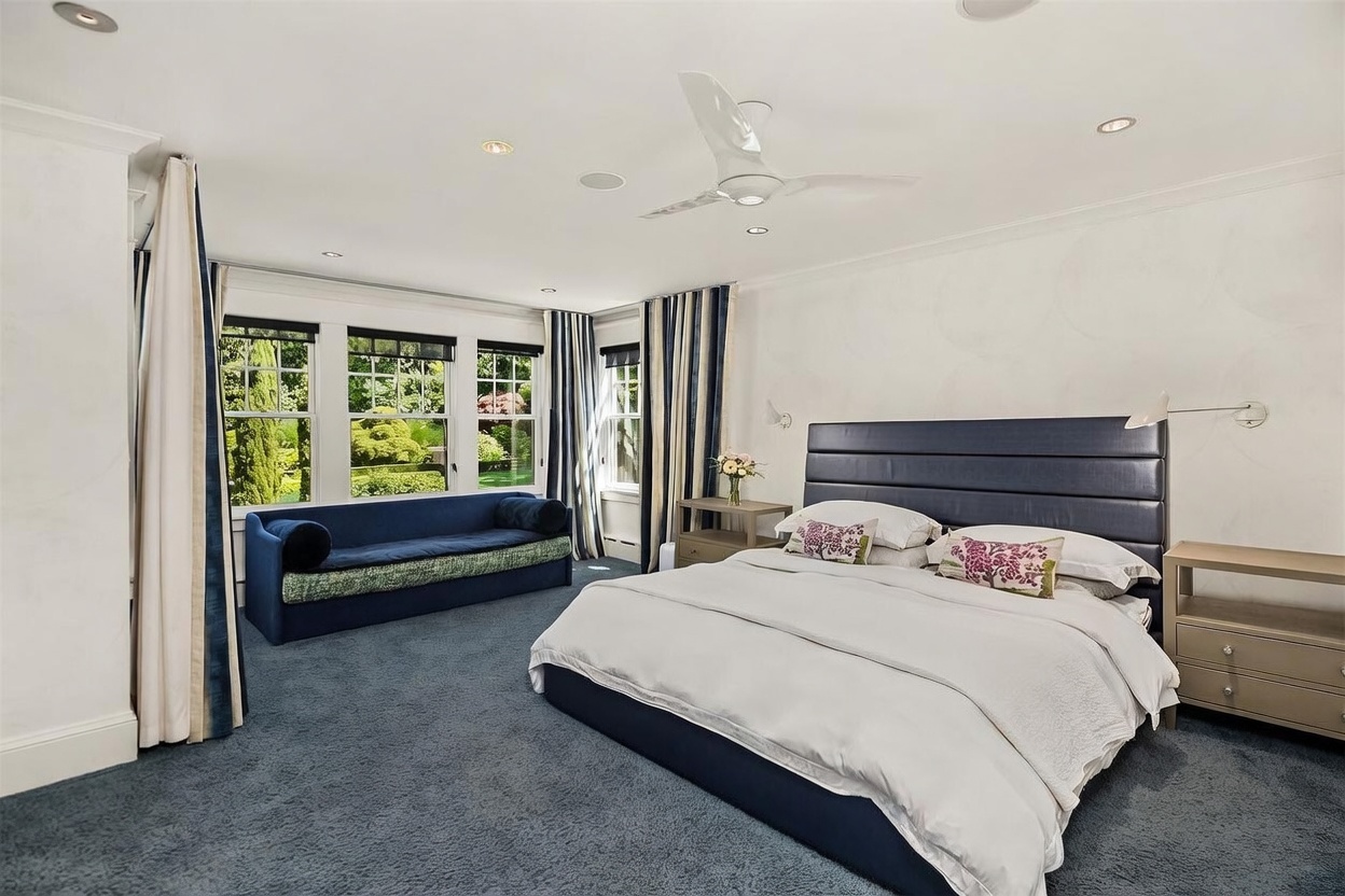 This spacious primary bedroom features a large, dark blue upholstered headboard and a matching blue daybed positioned beneath a bank of windows that overlook a lush green garden. The room is carpeted in a deep blue tone and includes two light-colored nightstands, a ceiling fan, and soft, neutral-toned walls. The perspective is from the corner of the room, capturing the expansive layout and the bright, natural light streaming in from the exterior.