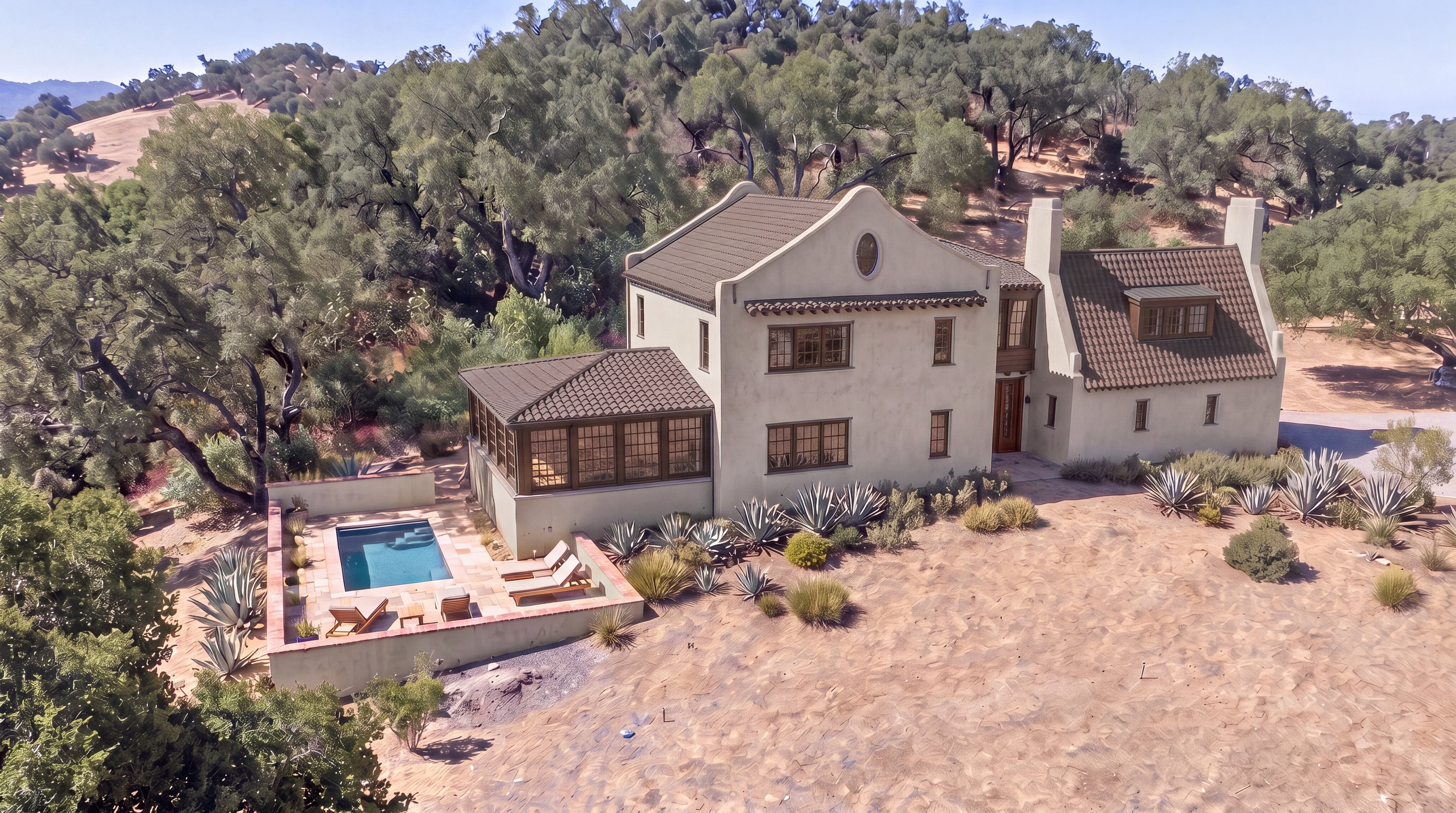 This stunning Mediterranean-style home is captured from an elevated perspective, showcasing its multi-level roofline, cream-colored stucco exterior, and charming sunroom addition. The property features a private swimming pool area set within a terraced landscape, surrounded by native desert flora and a backdrop of rolling hills. The combination of architectural elegance and secluded outdoor living spaces makes this a highly desirable residence.