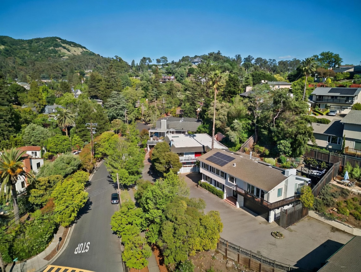 This high-angle aerial view captures a residential neighborhood nestled in a lush, hilly landscape. The scene highlights a multi-level home with a dark shingled roof equipped with solar panels, situated along a winding road with a visible stop sign. The surrounding area is densely wooded with mature trees, providing a sense of privacy and integration with the natural environment.
