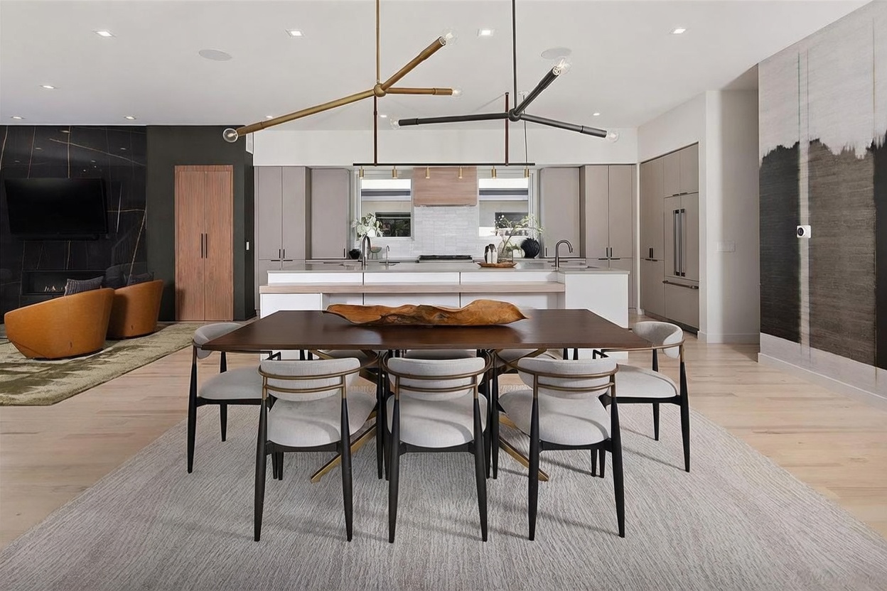 This open-concept space features a central dining area with a dark wood table surrounded by six upholstered chairs, set atop a textured area rug. In the background, a sleek kitchen with light-toned cabinetry and a large island serves as a focal point, while a living area with leather armchairs and a dark stone-clad wall is visible to the left. The room is illuminated by two striking, multi-armed modern light fixtures that hang above the dining table and kitchen island, creating a sophisticated and cohesive aesthetic.