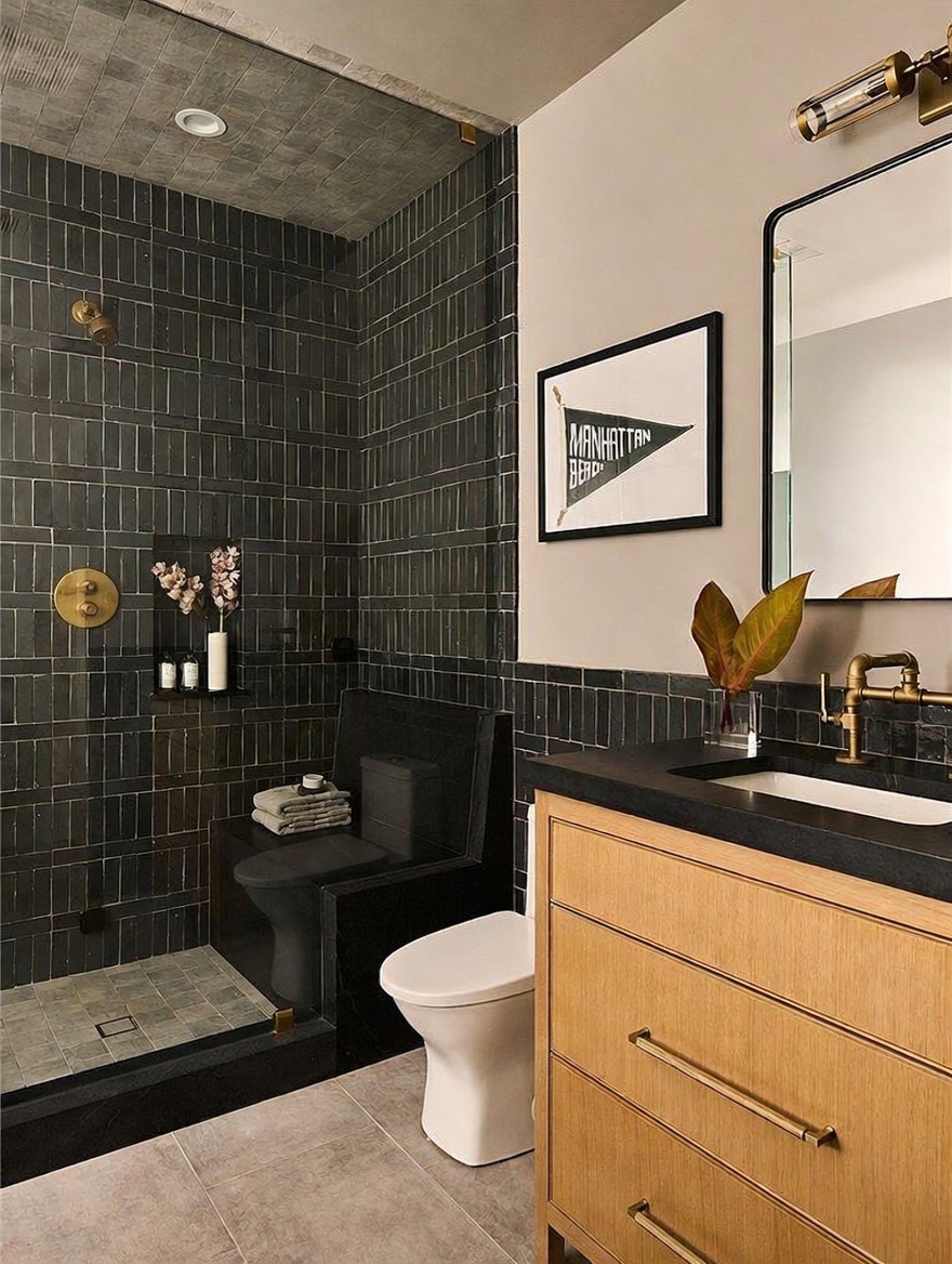 This modern bathroom features a striking walk-in shower clad in dark, vertical-stacked tiles that extend to the ceiling. A light wood vanity with a dark countertop and brass-toned fixtures provides a warm contrast, while a framed Manhattan Beach print adds a personalized touch. The space is completed by a white toilet and a large, rectangular mirror, creating a sophisticated and contemporary aesthetic.