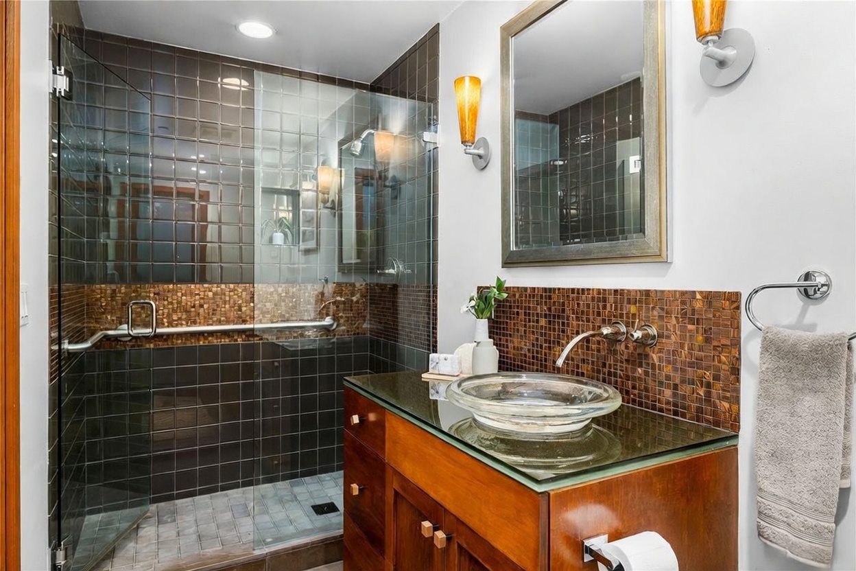 This modern bathroom features a striking glass vessel sink atop a rich wooden vanity, complemented by a mosaic tile backsplash and a walk-in shower with dark square tiles. The space is accented by warm, amber-toned wall sconces and a framed mirror, creating a sophisticated and spa-like atmosphere. The perspective is a medium shot capturing the vanity and shower area, highlighting the high-end finishes and textures.