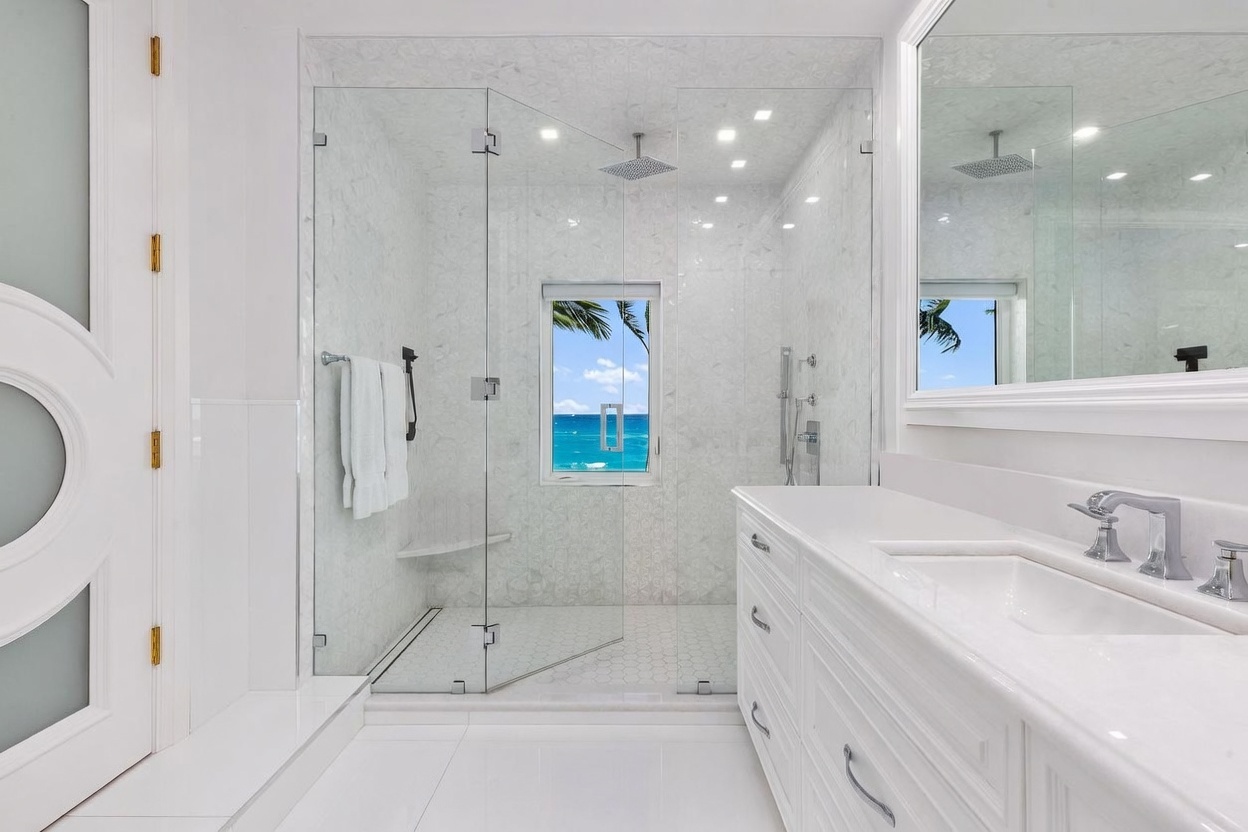This bright, modern bathroom features a spacious walk-in shower enclosed by glass panels, showcasing intricate patterned wall tile and a built-in bench. A long white vanity with a stone countertop and dual sinks sits opposite the shower, complemented by a large mirror reflecting the space. A window within the shower area provides a clear, scenic view of the ocean, adding a luxurious and airy feel to the room.