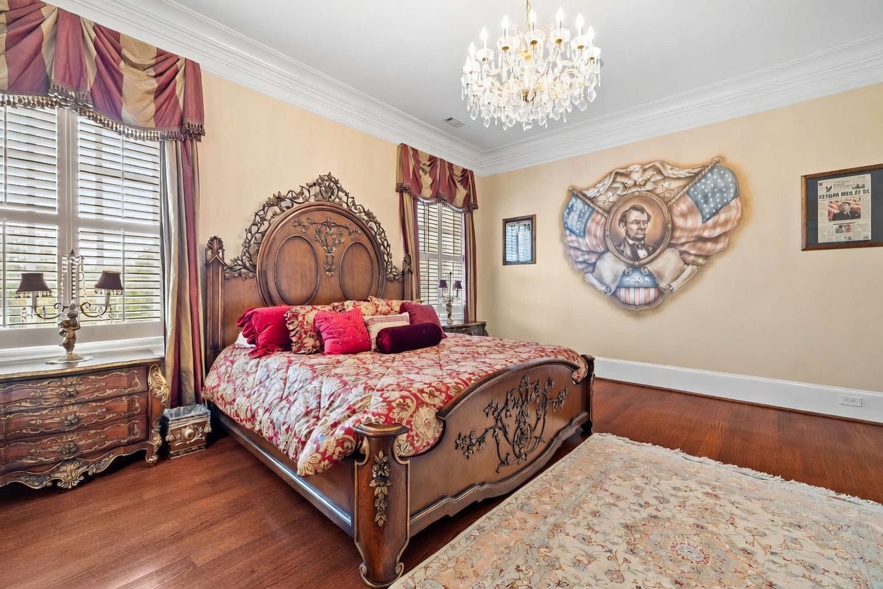 This primary bedroom features a traditional, ornate aesthetic with a large wooden bed frame as the centerpiece, dressed in patterned red and gold bedding. The room is accented by a prominent wall mural depicting Abraham Lincoln framed by American flags, and a crystal chandelier hangs from the ceiling. Large windows with plantation shutters and heavy fabric valances provide natural light, while a patterned area rug sits atop the hardwood flooring.