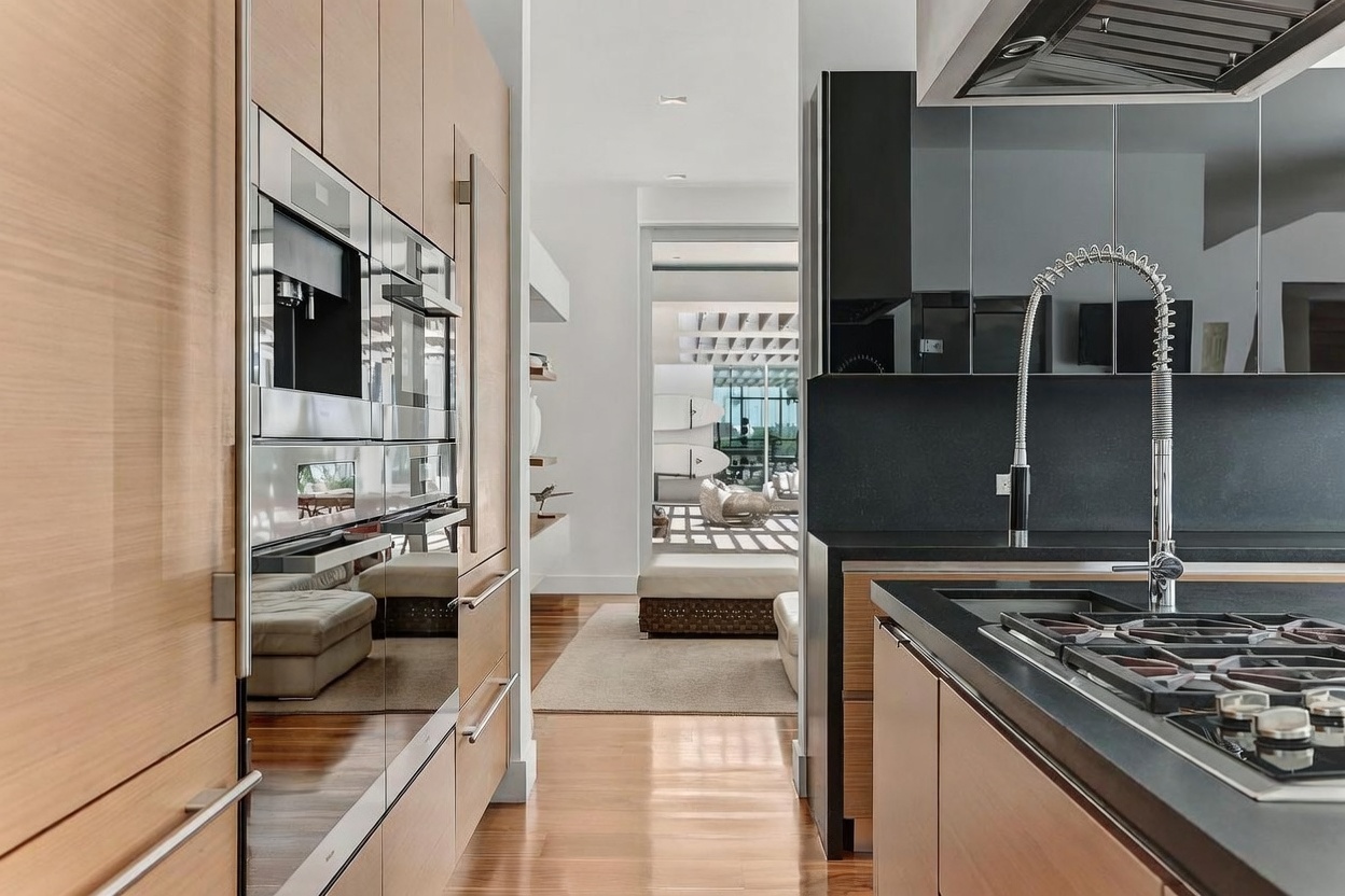 This modern kitchen features a sleek design with light wood cabinetry and dark, matte-finish countertops. The space includes integrated stainless steel appliances and a prominent gooseneck faucet over the cooktop, creating a sophisticated and functional culinary environment. A clear line of sight extends through the kitchen into an adjacent living area, emphasizing an open-concept layout with warm wood flooring throughout.
