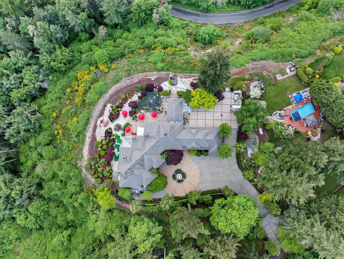 This high-angle aerial perspective captures a sprawling luxury estate nestled within a lush, wooded landscape. The property features a large, multi-winged residence with a circular driveway, a private swimming pool area, and meticulously landscaped gardens including a pond and a putting green. The expansive grounds are surrounded by dense forest, emphasizing privacy and a secluded, resort-like atmosphere.