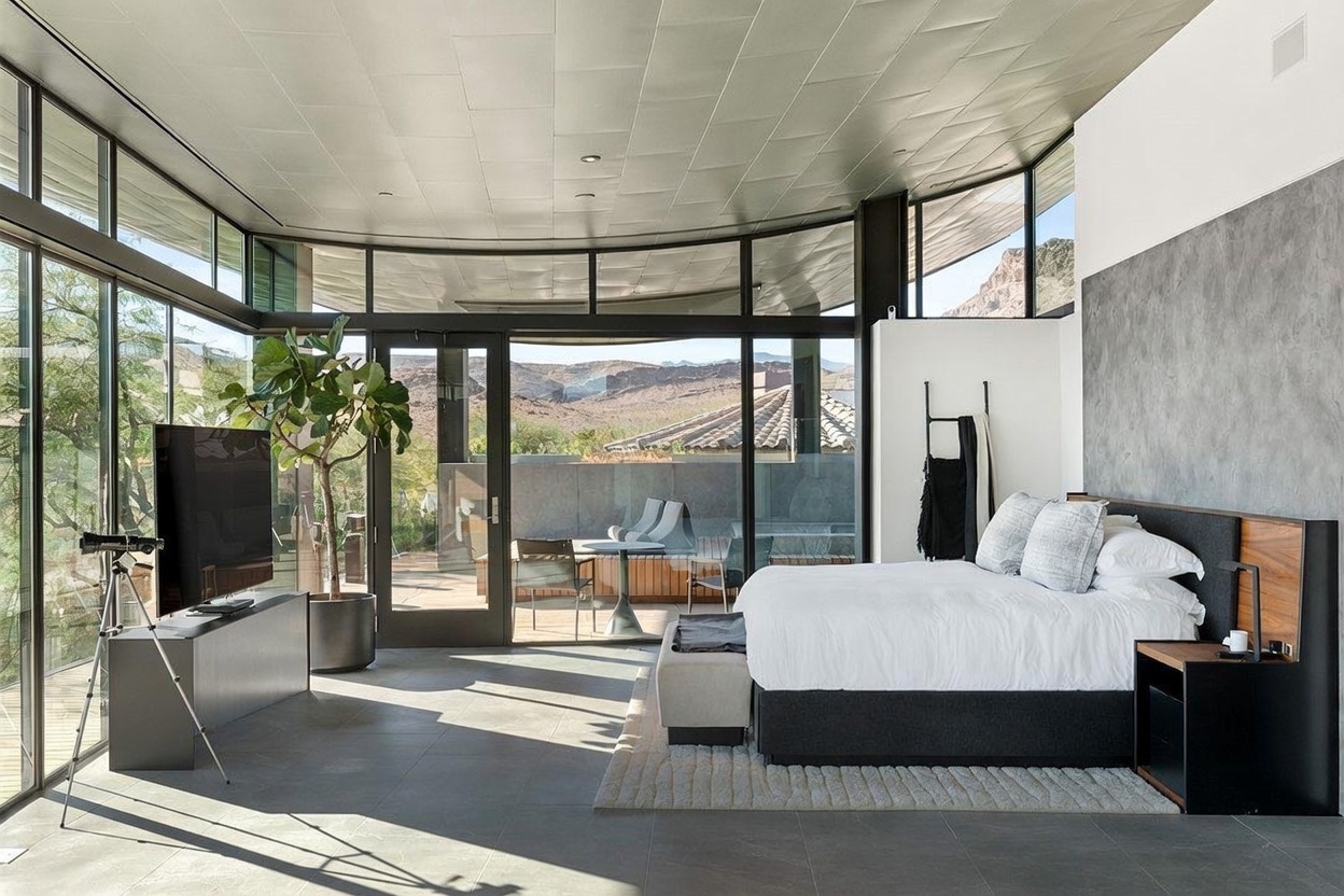 This modern primary bedroom features expansive floor-to-ceiling glass walls that provide panoramic views of the desert landscape and mountains. The room is anchored by a large bed with a dark headboard and a neutral-toned rug, while a sleek media console and a potted plant add to the minimalist aesthetic. A glass door opens directly onto a private patio, seamlessly blending indoor and outdoor living spaces.
