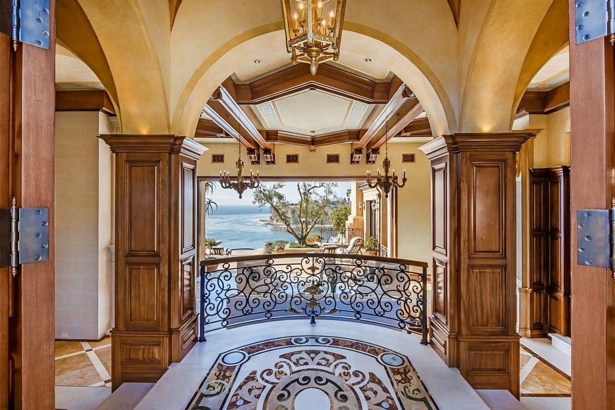 This grand entryway features a sophisticated foyer with an intricate, circular patterned stone floor inlay that leads toward a decorative wrought-iron railing. The space is framed by tall, arched openings with rich wood trim and columns, creating a dramatic transition into an open-air balcony overlooking the ocean. The perspective is centered, drawing the eye through the architectural archway toward the expansive coastal view beyond.