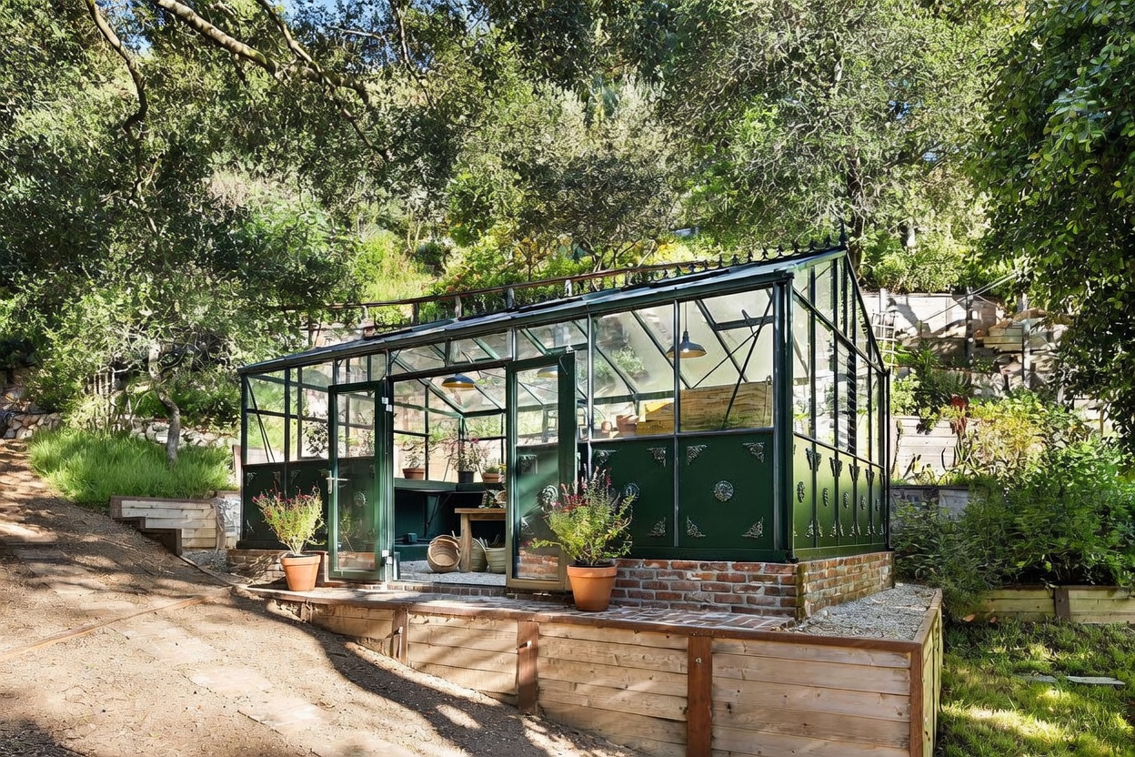This image features a charming, classic-style greenhouse set within a lush, sloped garden landscape. The structure is constructed with dark green metal framing and glass panels, resting on a raised brick foundation and a wooden retaining wall base. The surrounding area is filled with mature trees and greenery, creating a serene and private atmosphere perfect for gardening enthusiasts.