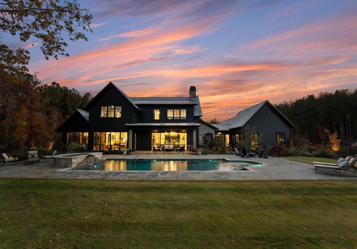 This stunning dark-sided modern farmhouse is captured at dusk, showcasing a sprawling rear elevation with a large swimming pool and stone patio in the foreground. The home features a mix of dark vertical siding and stone accents, with large windows that glow warmly against the vibrant sunset sky. The expansive lawn and surrounding mature trees create a serene, private atmosphere perfect for outdoor entertaining.