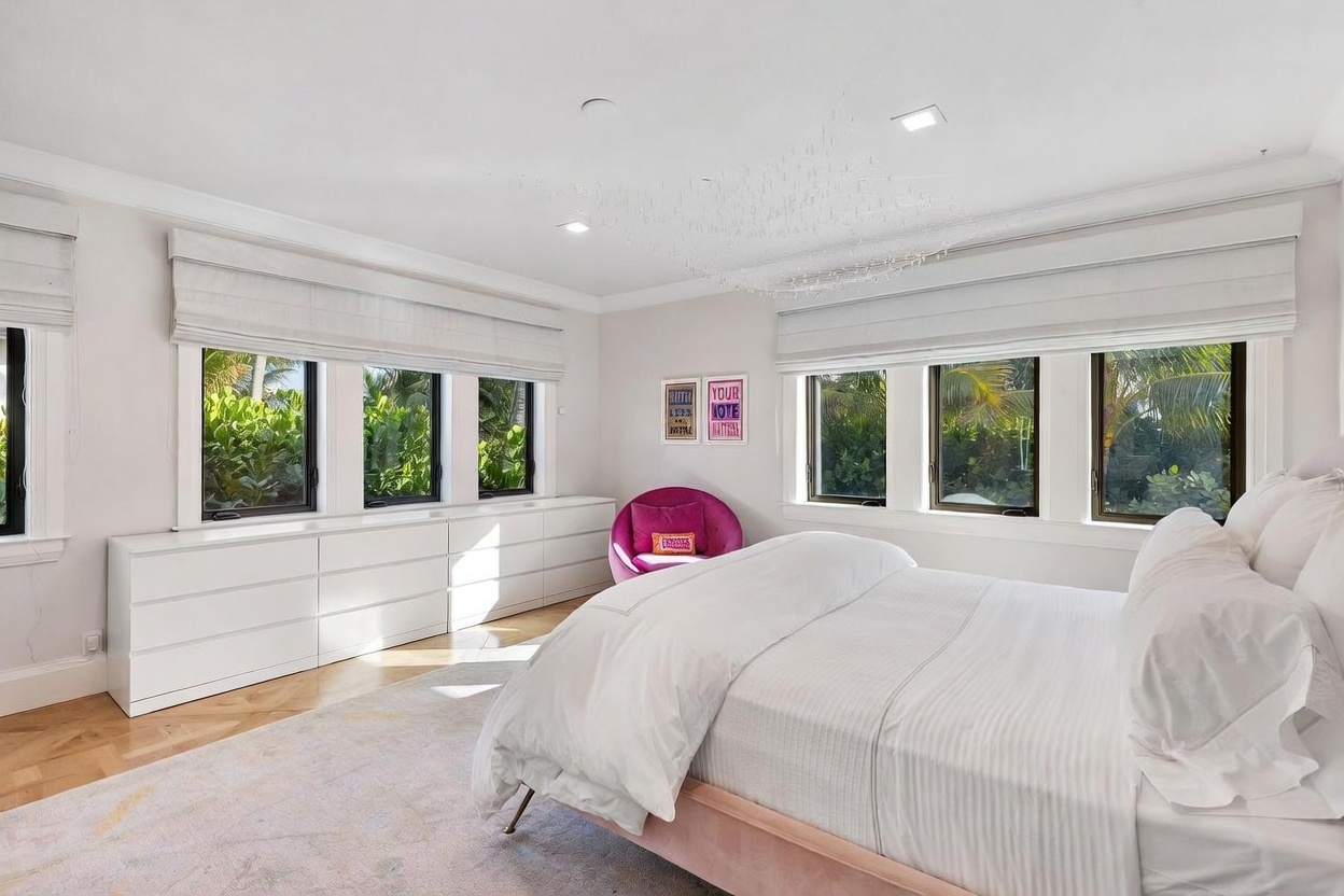 This bright and airy primary bedroom features a large bed with white linens as the central focus, complemented by a vibrant pink accent chair in the corner. A long, low-profile white storage unit runs beneath a series of windows, providing ample surface space and storage. The room is finished with light-toned wood flooring, a large area rug, and a unique, delicate chandelier, creating a clean and inviting atmosphere.