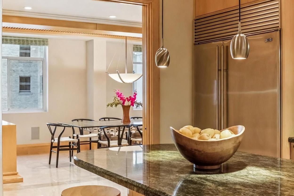 This view captures a sophisticated kitchen area featuring a polished stone-topped island with a wooden bowl of fruit, positioned in the foreground. Beyond the island, a doorway leads into a bright dining room furnished with a glass-topped table and modern chairs, accented by a floral centerpiece. The kitchen is equipped with a large stainless steel refrigerator and elegant pendant lighting, creating a seamless and open transition between the two living spaces.