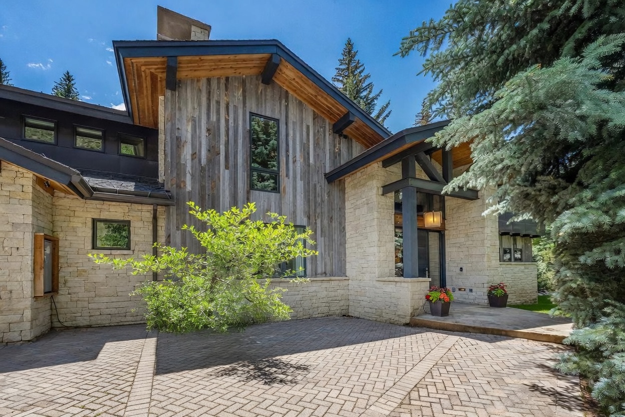 This striking mountain-style home features a sophisticated blend of light-toned stone masonry and vertical weathered wood siding. The entryway is framed by dark structural beams and a covered porch, leading to a paved walkway surrounded by lush landscaping and vibrant purple flowers. The perspective captures the home's architectural depth and natural integration with the surrounding evergreen trees.