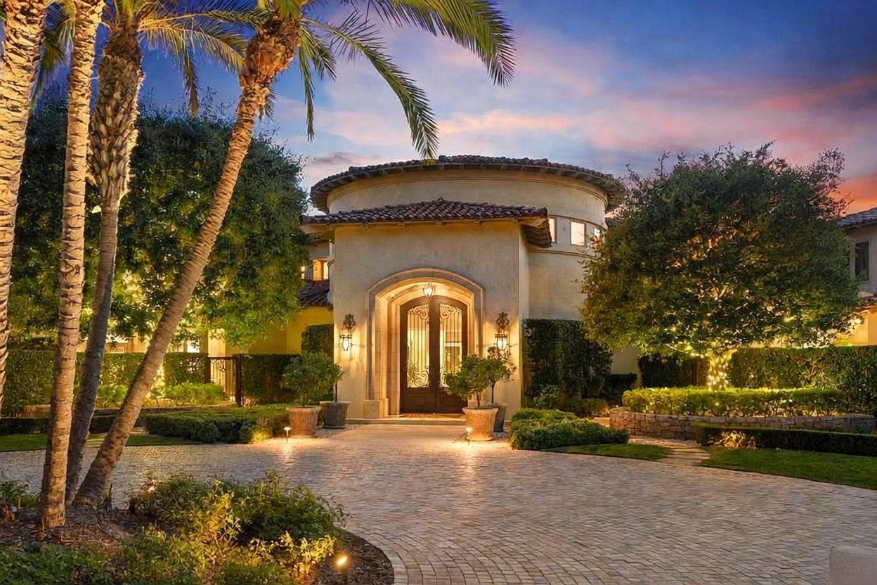 This is a front exterior view of a luxurious home at dusk, showcasing a circular entryway with an arched double door and decorative sconces. The property features meticulously landscaped grounds with mature trees, manicured hedges, and a circular driveway paved with brick. The warm lighting highlights the architectural details and creates an inviting ambiance.