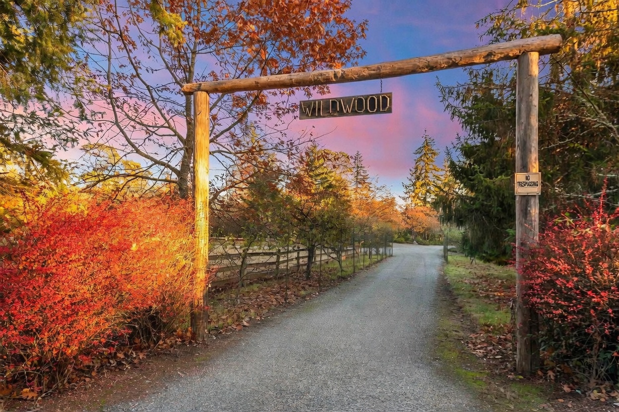 This image captures the rustic entrance to a property, marked by a large wooden archway featuring a sign that reads 'WILDWOOD.' A gravel driveway leads through the gate, flanked by vibrant autumn foliage and a wooden fence, under a dramatic sunset sky. The perspective is a ground-level shot looking down the path, emphasizing the welcoming and secluded nature of the property.