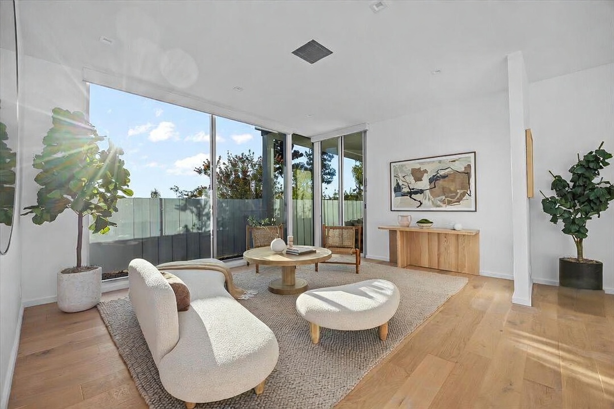 This bright and airy living room features light wood flooring and a neutral color palette, creating a serene and modern atmosphere. The space is anchored by a textured area rug, a curved sofa, and a round wooden coffee table, all positioned to take advantage of the large windows that offer views of the outdoor area. A console table sits against the wall beneath a piece of abstract art, while two potted plants add a touch of greenery to the room.